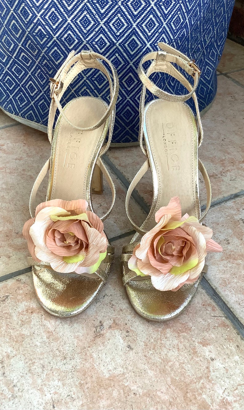 May include: A pair of gold sandals with a flower embellishment on each strap. The sandals are labeled "Office London".