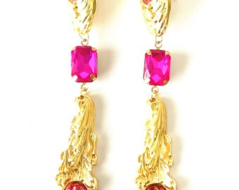 May include: A pair of gold tone dangle earrings with pink crystal accents. The earrings feature a floral design and three pink crystals of different shapes.