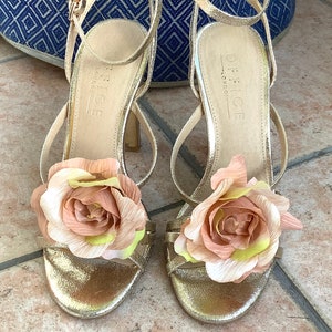 May include: A pair of gold sandals with a flower embellishment on each strap. The sandals are labeled "Office London".