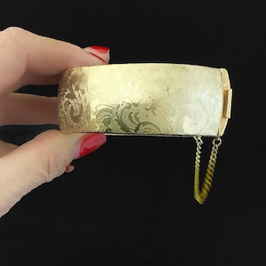 May include: A gold-coloured bangle bracelet with an engraved floral pattern. The hinged bracelet has a safety chain and a rectangular shape. The bracelet is held against a black background.