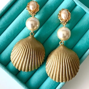 Puede incluir: Un par de pendientes de concha dorada con detalles de perlas blancas. Los pendientes se muestran en una caja de joyería turquesa.