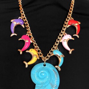 May include: Gold-tone chain necklace with colourful dolphin charms in purple, pink, yellow, red, and blue. A large, turquoise seashell pendant hangs at the centre. A statement necklace.