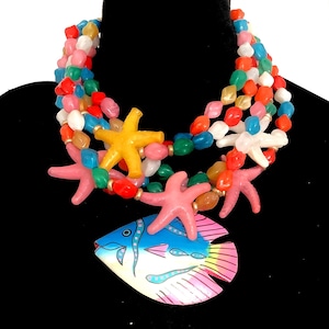 May include: A colorful necklace featuring multiple strands of multi-colored beads. The necklace is adorned with starfish and a fish pendant. The fish pendant is blue, pink, and yellow. The starfish are yellow, pink, and white.