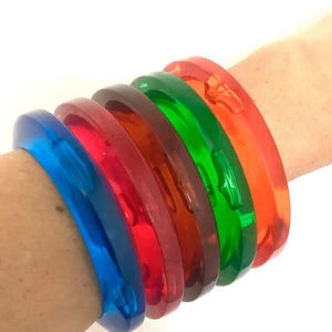 May include: A collection of six translucent, colorful bangles in shades of blue, red, brown, green, and orange. The bracelets are stacked on a wrist, showcasing their vibrant hues and glossy finish. These accessories are perfect for adding a pop of color.