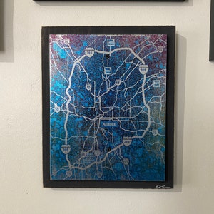 May include: A framed map of Atlanta, Georgia with a gradient background of pink, blue, and purple. The map is printed on a silver metallic surface and shows major highways and interstates. The frame is black and the map is signed in the bottom right corner.
