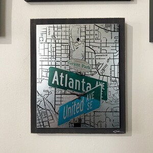 May include: A silver and black framed artwork featuring a map of Atlanta, Georgia with three street signs that read "Grant Park", "Atlanta Ave", and "United Ave SE".