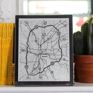 May include: Black and white map of Atlanta, Georgia, with highway numbers and the city name printed in the center. The map is framed in a black frame and is displayed on a shelf with a cactus and a stack of yellow National Geographic magazines.