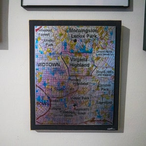 May include: A colorful map of Atlanta, Georgia, with neighborhood names highlighted in black text. The map is framed in a black frame with a gold trim.