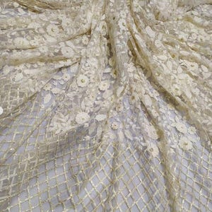 May include: Cream-coloured fabric with a floral pattern and a diamond grid design. The fabric features raised floral embroidery with small, gold-coloured embellishments. The material is draped, showing the texture and detail of the design.