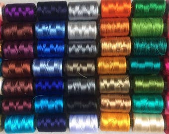 Könnte beinhalten: Eine Sammlung bunter Garnrollen, in ordentlichen Reihen angeordnet. Die Fäden sind in verschiedenen Farben erhältlich, darunter Rosa, Orange, Grün, Blau und Braun. Die Spulen sind fest gewickelt und scheinen aus einem glatten, glänzenden Material zu bestehen. Ideal für Näh- und Bastelprojekte.