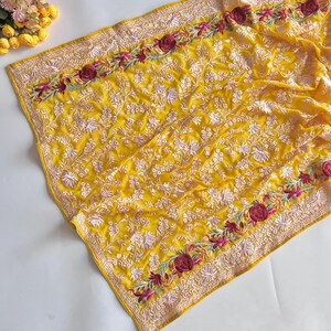 Pode incluir: Um tecido amarelo brilhante com um intrincado bordado floral branco em toda a superfície e uma borda decorativa com motivos de rosas em vermelho escuro e verde. O têxtil apresenta um padrão denso de pequenas flores e folhas brancas.