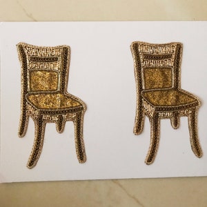 May include: Two gold beaded chair appliques with a brown outline. The chairs have a simple design with a back, seat, and four legs.