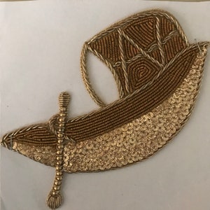 May include: A gold-colored, embroidered boat-shaped applique. The boat features a detailed sail, a sequined lower section, and a decorative oar. The applique is likely for embellishing clothing or accessories.