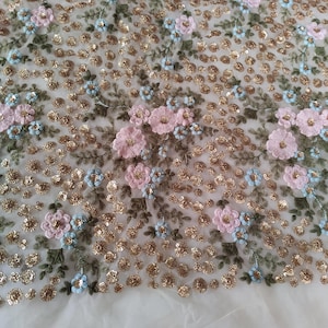 May include: Close-up of a sheer fabric with intricate floral embroidery. The design features pink and light blue flowers, green leaves, and gold sequins. The delicate details create a textured, decorative surface, suitable for apparel or craft projects.