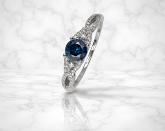 Könnte beinhalten: Ein silberner Ring mit einem großen, runden, tiefblauen Edelstein als Mittelpunkt. Das Band weist ein kompliziertes, ineinander verwobenes Design auf, das mit kleineren, klaren Edelsteinen verziert ist. Der Ring ist vor einem weißen Marmorhintergrund platziert.