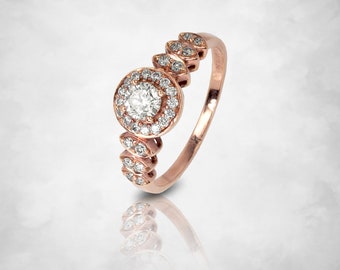 Può includere: Anello in oro rosa con un diamante rotondo centrale circondato da un alone di diamanti più piccoli. La fascia è impreziosita da dettagli di diamanti, creando un design scintillante ed elegante. L'anello è presentato su uno sfondo morbido e chiaro.