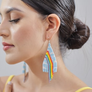 May include: A woman wearing a pair of long, colorful beaded earrings. The earrings are blue, white, red, orange, yellow, green, and purple. The earrings are shaped like a teardrop and have a fringe at the bottom.