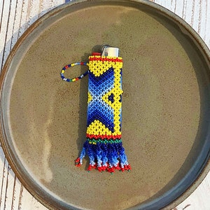 May include: A beaded lighter cover with a blue, yellow, and red geometric pattern. The cover has a tassel fringe and is sitting on a brown plate.