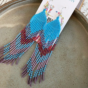 May include: A pair of long, beaded earrings with a gradient of turquoise, purple, and red beads. The earrings are hanging from a silver hook and are labeled "You're a Beauty".