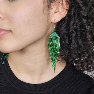 Op de afbeelding: Groene kralen oorbellen met een driehoekige bovenkant en bungelende strengen. De oorbellen hebben een levendige groene tint en lijken handgemaakt te zijn. De oorbellen worden door een persoon gedragen.