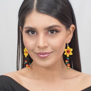 May include: A woman wearing yellow flower earrings with red and green accents. The earrings are dangling and have a beaded design. The woman is wearing a black off-the-shoulder top.