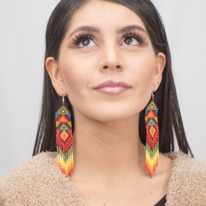 Puede incluir: Una mujer con el pelo largo y oscuro lleva un cárdigan marrón peludo y pendientes largos de cuentas de colores. Los pendientes son rojos, naranjas, amarillos, verdes, azules y negros.