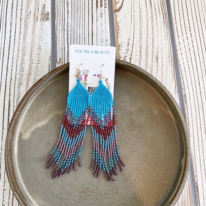May include: A pair of turquoise and purple beaded dangle earrings with a silver hook. The earrings are on a white card with the text "You're a Beauty".