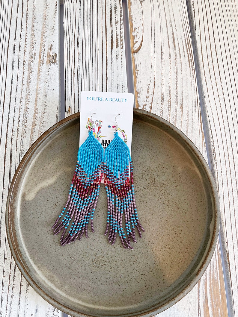 May include: A pair of turquoise and purple beaded dangle earrings with a silver hook. The earrings are on a white card with the text "You're a Beauty".