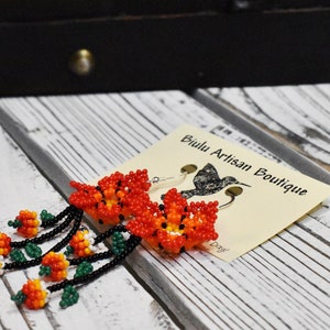 May include: Beaded earrings with a floral design in shades of orange, red, and green. The earrings are hanging from silver hooks and are labeled "Biulu Artisan Boutique".