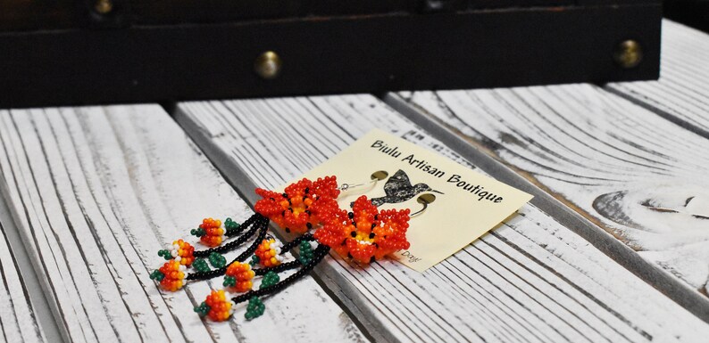 May include: Beaded earrings with a floral design in shades of orange, red, and green. The earrings are hanging from silver hooks and are labeled "Biulu Artisan Boutique".
