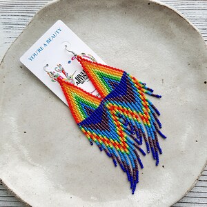 May include: Rainbow beaded earrings with a fringe design. The earrings are made with small, colorful beads and have a silver hook closure. The earrings are displayed on a white plate with the text "You're a Beauty" on a card.