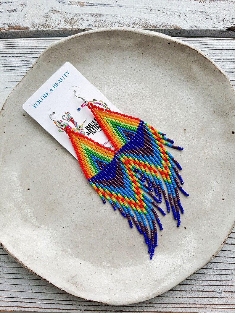 May include: Rainbow beaded earrings with a fringe design. The earrings are made with small, colorful beads and have a silver hook closure. The earrings are displayed on a white plate with the text "You're a Beauty" on a card.