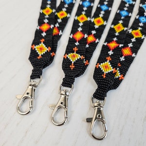 Peut inclure: Gros plan sur trois lanières noires ornées de motifs géométriques colorés. Les motifs présentent des losanges bleus, jaunes, oranges et blancs. Chaque lanière est dotée d'un mousqueton en métal argenté.