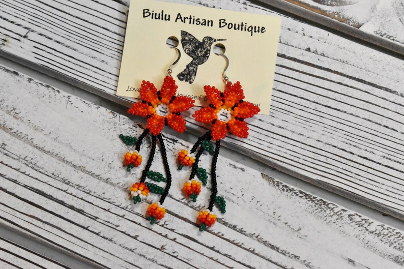 May include: A pair of orange and black beaded flower earrings with green leaves. The earrings are handmade and feature a delicate floral design. The earrings are hanging from silver hooks.