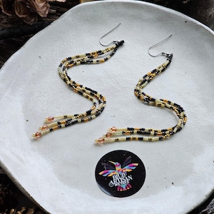 May include: A pair of long, beaded earrings with black, gold, and cream coloured beads. The earrings are hanging on a white plate with a sticker that says "BiJu Artisan Boutique".