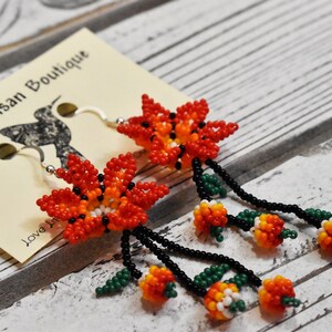 May include: A pair of beaded earrings featuring orange, red, and white flowers with green leaves and black beads. The earrings are hanging from silver hooks.