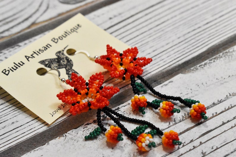 May include: A pair of beaded earrings featuring orange, red, and white flowers with green leaves and black beads. The earrings are hanging from silver hooks.