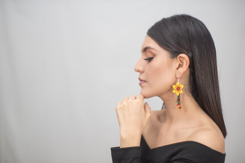 May include: A pair of vibrant, handmade earrings featuring a yellow flower design with red and green accents. The earrings dangle with beaded strands in shades of red, green, and yellow, creating a striking floral accessory.