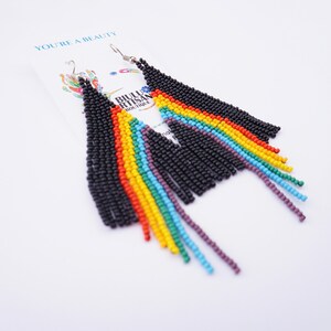 May include: A pair of beaded earrings with a black top section and rainbow fringe. The earrings are on a white card with the text "YOU'RE A BEAUTY" and the logo "BIJOU ARTISAN BOUTIQUE".
