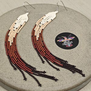 Può includere: Un paio di lunghi orecchini con perline con un gradiente di colori marrone, beige e bianco. Gli orecchini sono realizzati con piccole perline rotonde e hanno un design a frangia nella parte inferiore.