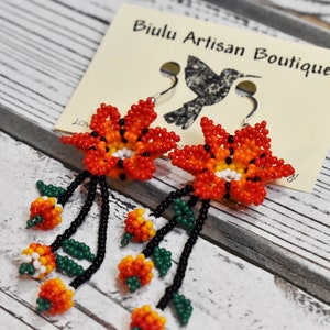 May include: A pair of beaded earrings featuring orange, red, yellow, white, and green flowers with black beads. The earrings are hanging from silver hooks and are labeled "Biulu Artisan Boutique".