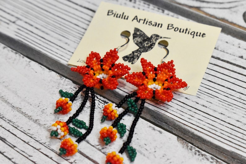 May include: A pair of beaded earrings featuring orange, red, yellow, white, and green flowers with black beads. The earrings are hanging from silver hooks and are labeled "Biulu Artisan Boutique".