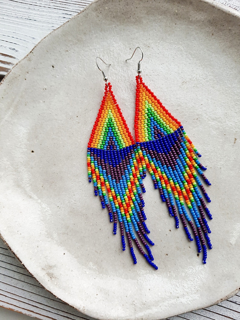 May include: A pair of rainbow beaded dangle earrings with a fringe of colorful beads. The earrings are made with small seed beads in a rainbow pattern, with a blue and purple fringe.