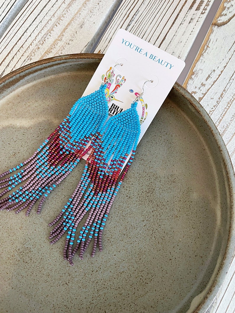 May include: A pair of long, beaded earrings with a gradient of turquoise, purple, and red beads. The earrings are hanging from a silver hook and are labeled "You're a Beauty".