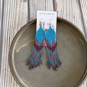 May include: A pair of long, beaded earrings with a gradient of turquoise, purple, and red beads. The earrings are hanging from a card that says "You're a Beauty".