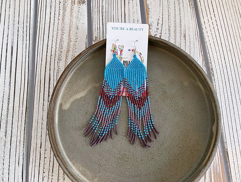 May include: A pair of long, beaded earrings with a gradient of turquoise, purple, and red beads. The earrings are hanging from a card that says "You're a Beauty".