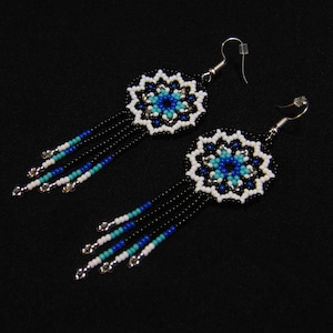 May include: A pair of beaded earrings with a floral design. The earrings feature a central flower in shades of blue, surrounded by white beads. Black beads form the fringe, accented with turquoise, blue, and silver beads. The earrings hang from silver-coloured hooks.