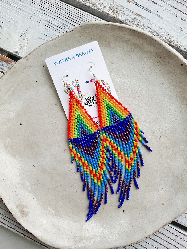 May include: Rainbow beaded earrings with a star design and a fringe. The earrings are on a white plate with the text "You're a Beauty" on a card.
