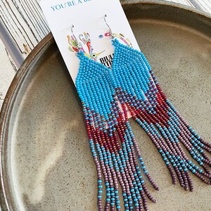 May include: A pair of long, beaded earrings with a turquoise, red, and purple color scheme. The earrings are hanging from a white card with the text "You're a Beauty".