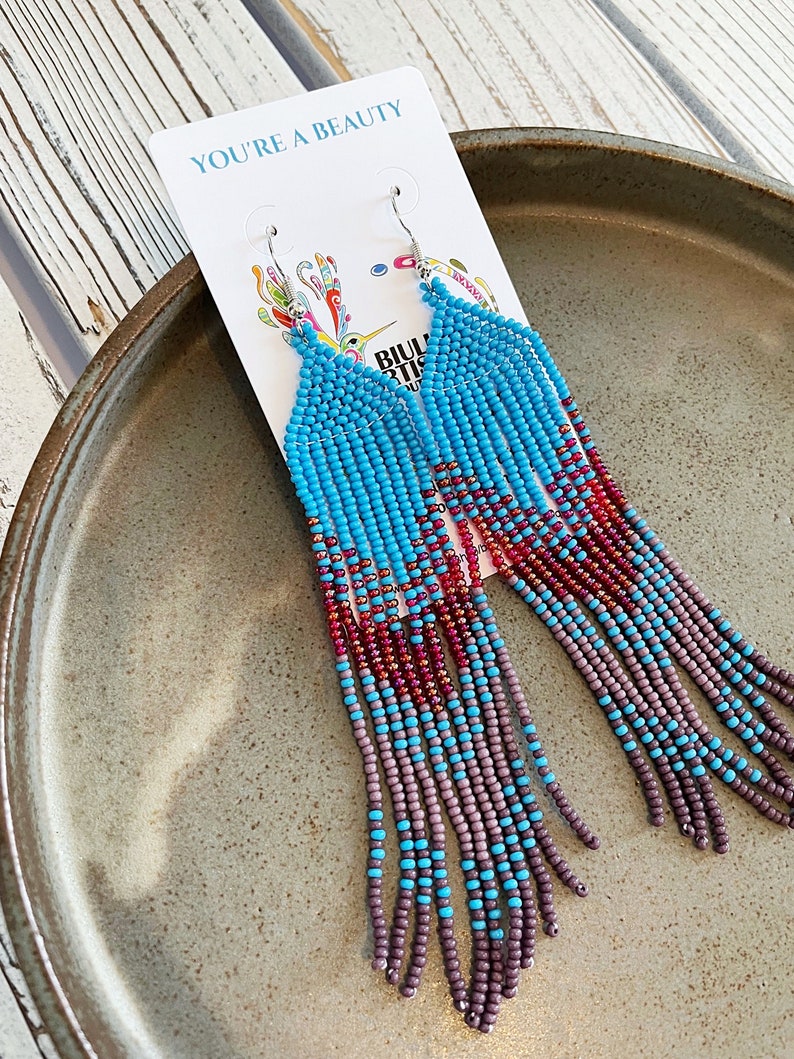 May include: A pair of long, beaded earrings with a turquoise, red, and purple color scheme. The earrings are hanging from a white card with the text "You're a Beauty".